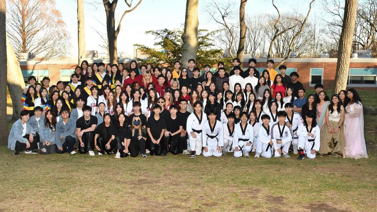 Around a hundred students posed in deep rows on grass outside a long brick school building, bare winter trees behind them. Some wear white martial-arts uniforms with black belts, others sashes or long formal dress.