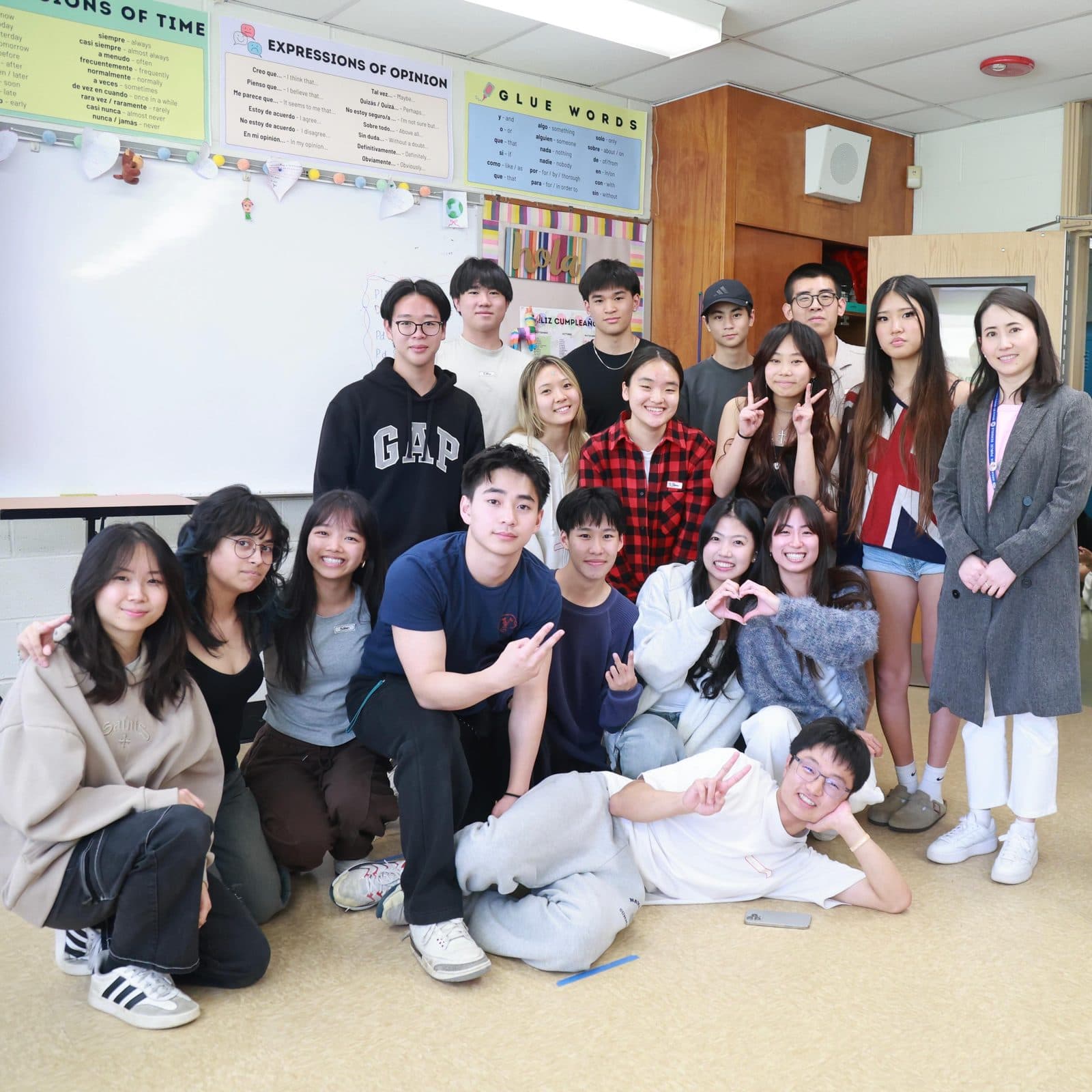 About seventeen students crowded together in a classroom in front of a whiteboard hung with Spanish grammar posters. Many make peace signs or heart shapes with their hands, and one lies stretched across the floor at the front.