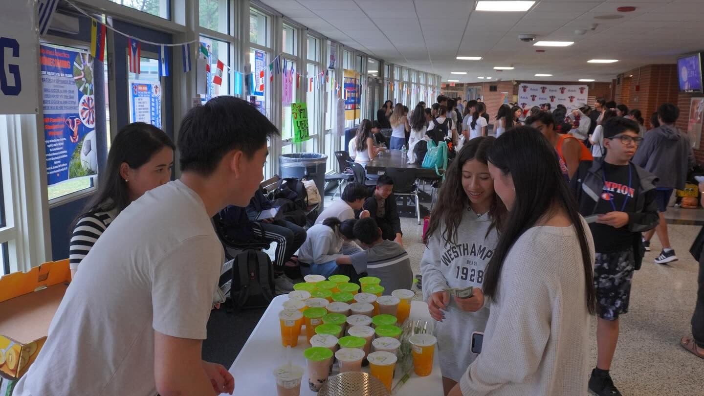 A long table in a busy school hallway set with rows of lidded plastic cups of orange drink and bubble tea. Two students stand behind it serving; two more in front are handing over cash, with a crowded corridor stretching away behind them.