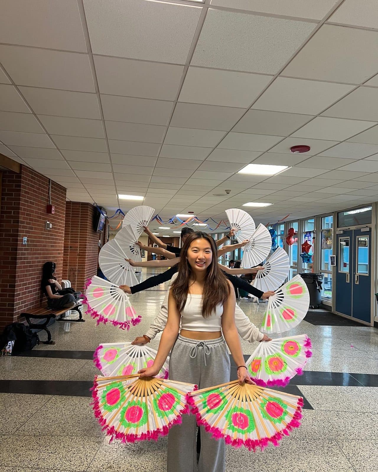 A student stands in a school corridor holding a large hand-painted paper fan open in each hand, the fans trimmed with pink feathers. Behind her several more dancers are lined up with their own fans raised and spread wide.