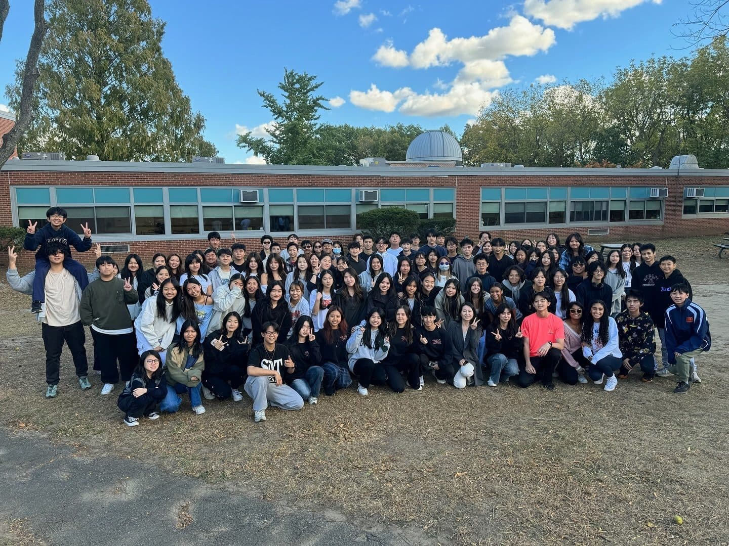 Around a hundred students packed into rows on the grass in front of a low brick school building under a blue sky with scattered cloud. Many make peace signs; at the left one sits up on another's shoulders.