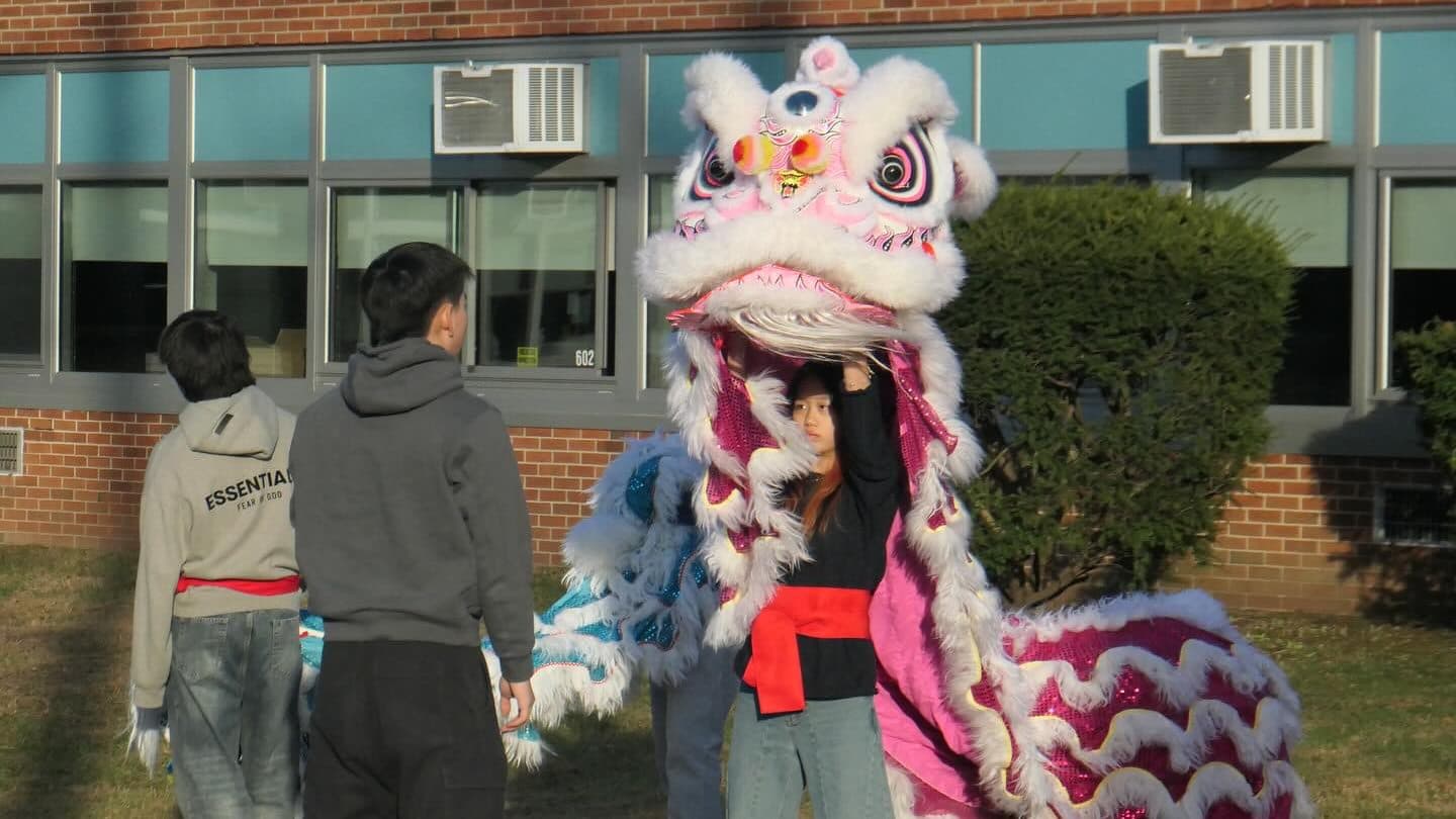 A student holds the head of a pink and white lion dance costume above her own head on grass outside a low brick school building. Two other students stand to the left with their backs to the camera, carrying a second costume.