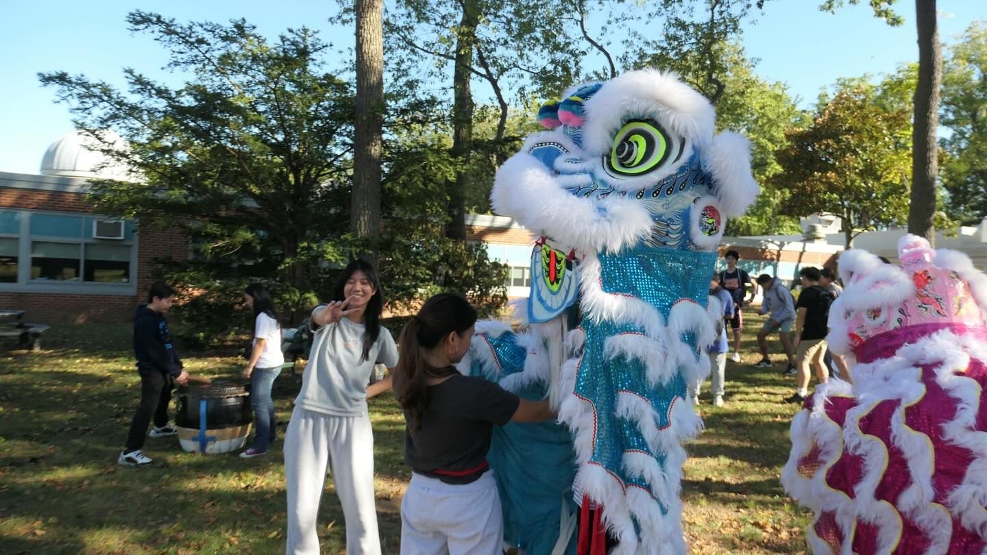 Students on a sunlit lawn handling two lion dance costumes. A blue and silver lion is held upright at the centre while a pink and white one crowds the right edge of the frame; a student at the left grins at the camera making a peace sign.