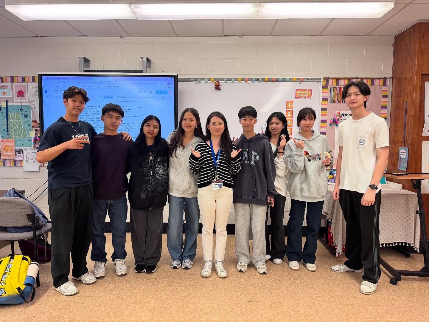 Nine students and an adult stand shoulder to shoulder in a single row across a classroom, a lit smartboard and a whiteboard behind them. Several make small heart signs with their fingers.
