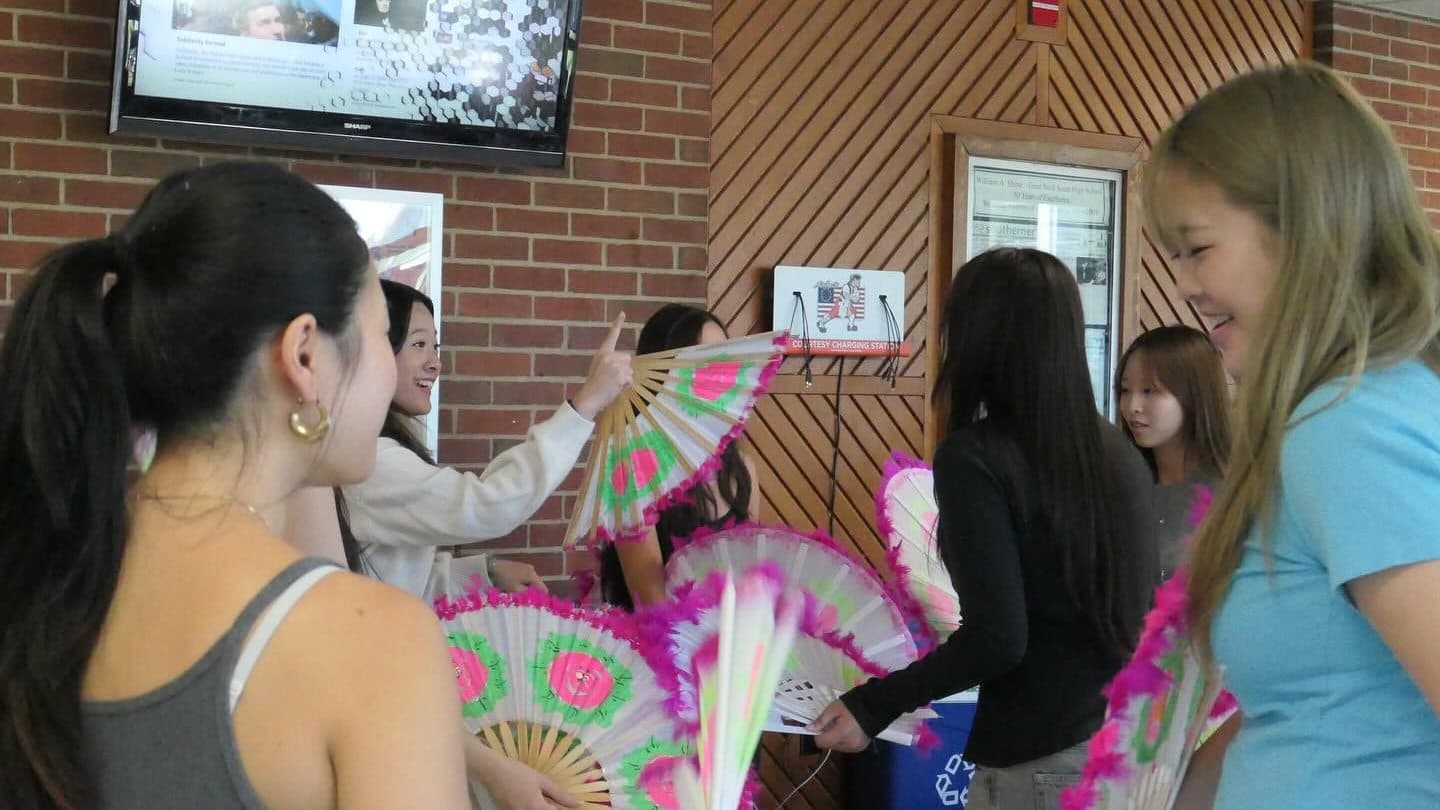 Students gathered around an indoor table holding large hand-painted fans edged in bright pink feathers. One reaches up to point at a small sign on the brick wall while the others look on, laughing.