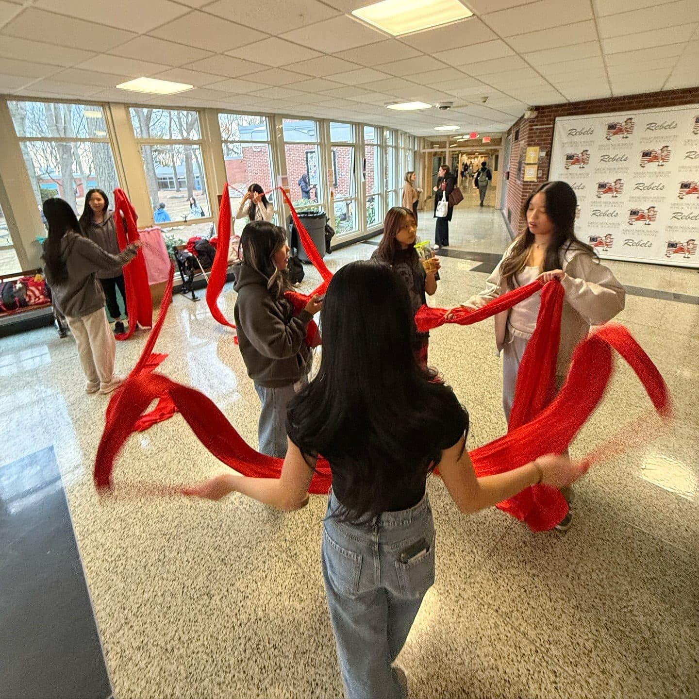 Students stand in a loose ring on a polished school lobby floor, each sweeping a long red ribbon through the air so the ribbons arc and loop between them. One dancer is seen from behind in the foreground.