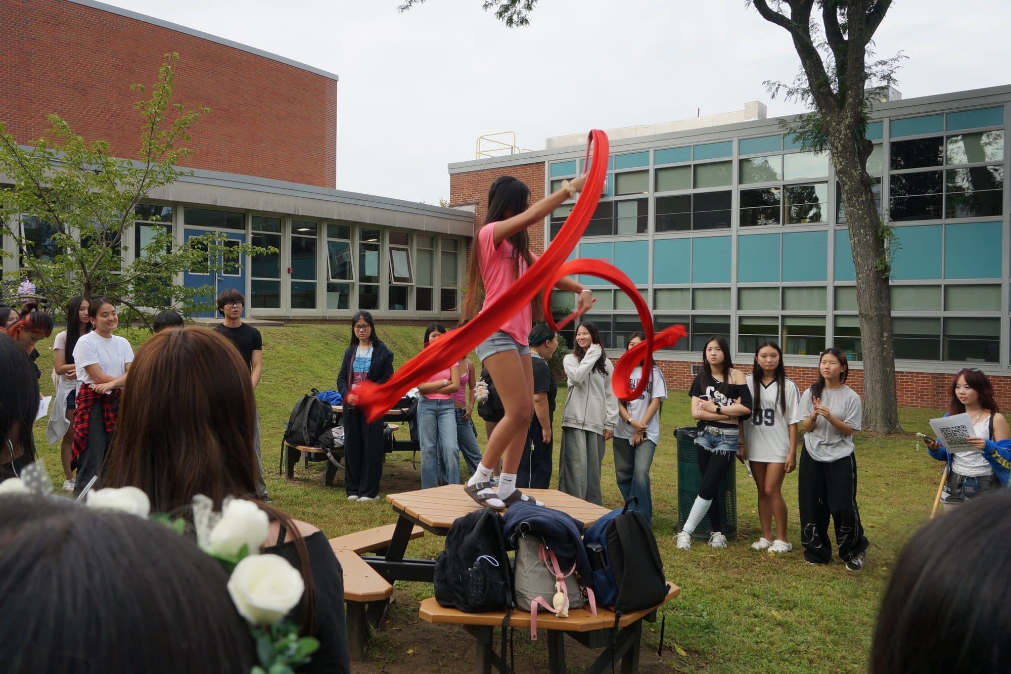 A student balances on a picnic table in a grassy school courtyard, sweeping a long red ribbon into a loop above her head. Students stand watching in a ring around her, with the school's brick and glass frontage behind.