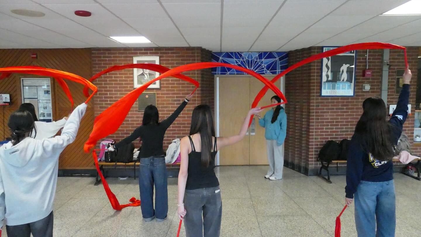 Five students stand well apart in a school lobby with their backs to the camera, each whirling a long red ribbon overhead so the ribbons draw wide loops across the brick wall. Another student faces them from across the floor.
