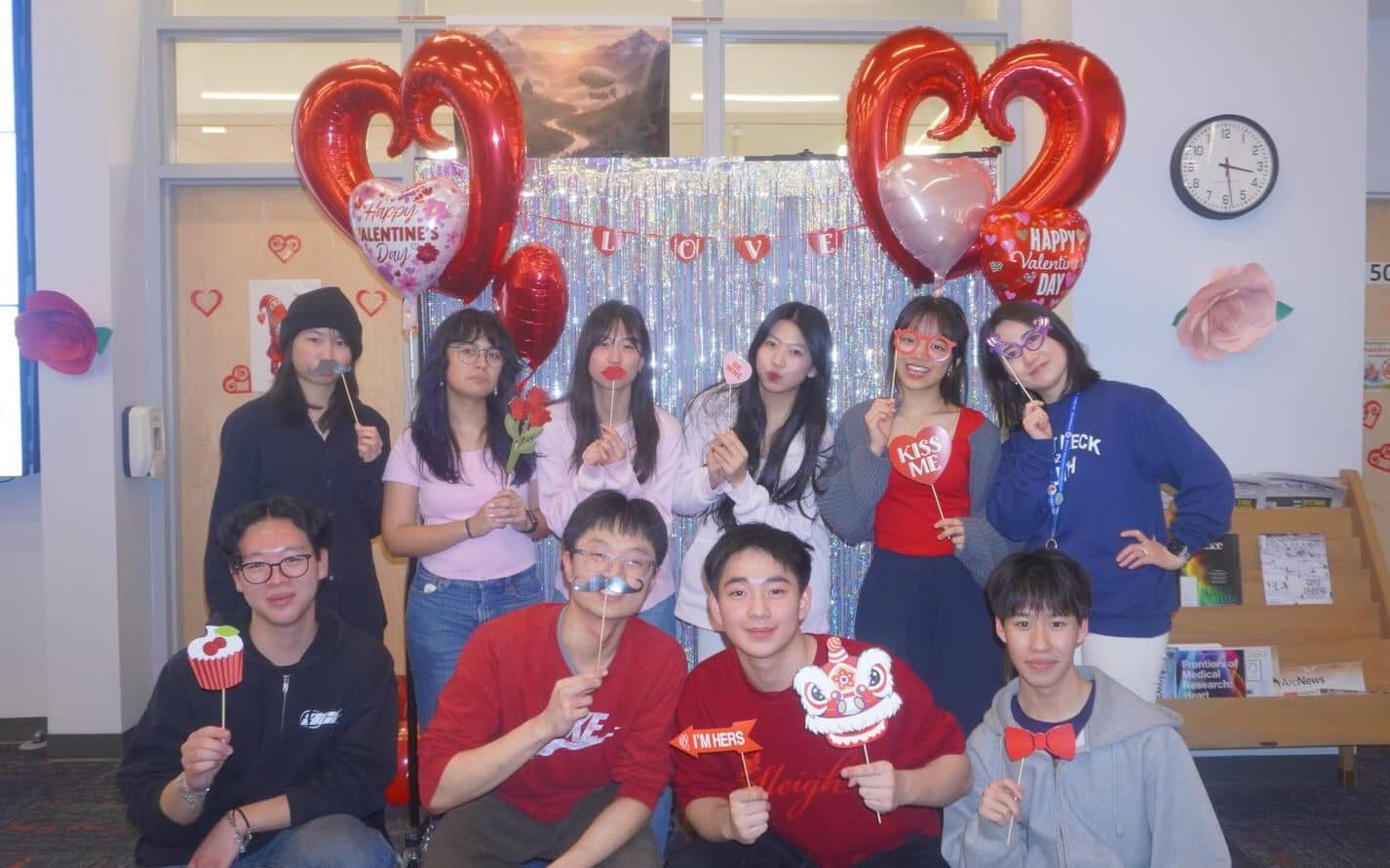 Eleven students posed in two rows in front of a silver fringe curtain hung with a LOVE banner, red heart balloons floating at either side. Each holds a paper photo-booth prop on a stick — lips, a moustache, glasses, a small lion's head.